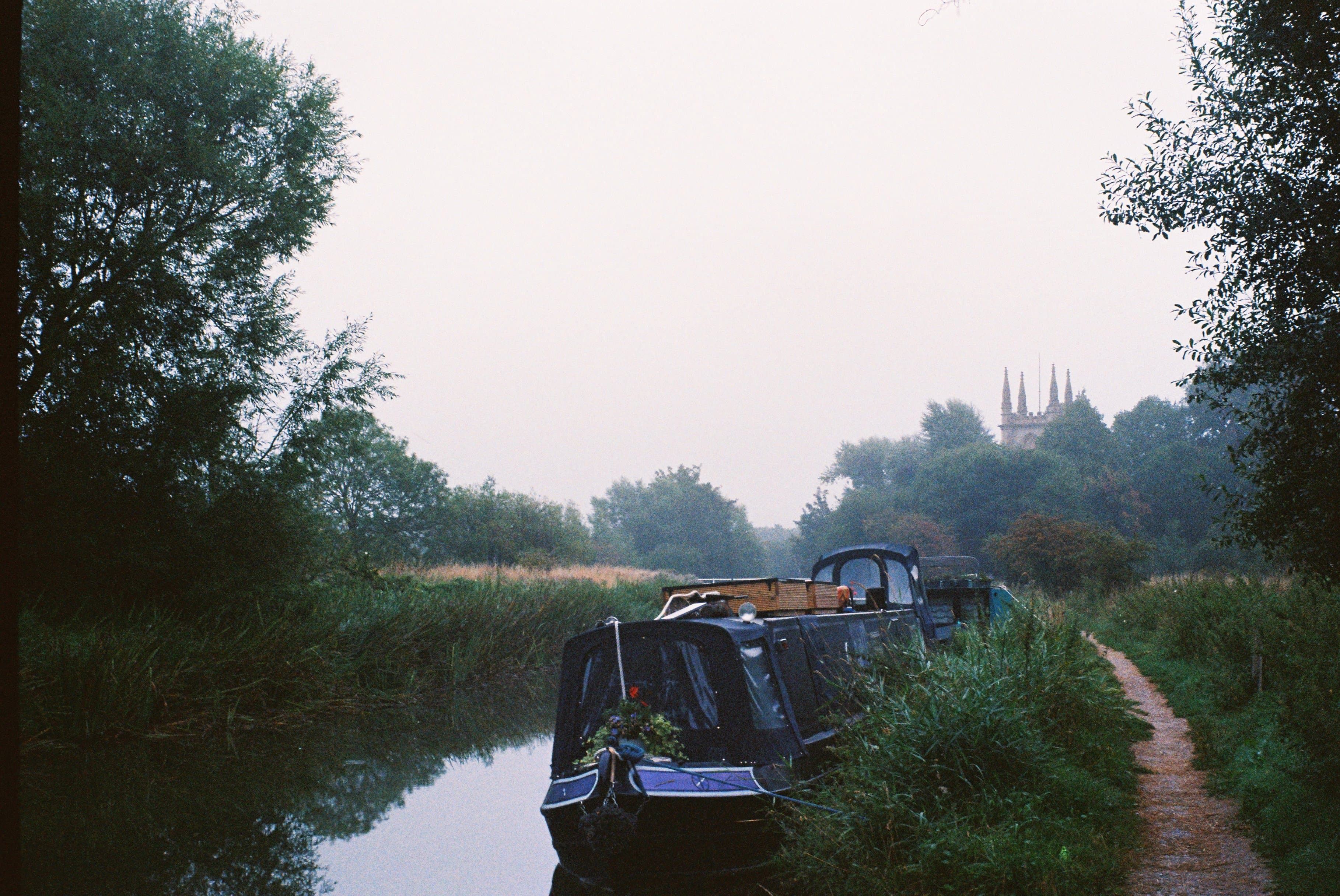 A narrow water canal. The rear end of a canal boat is visible and a dirt footpath is seen on the right side of the image. In the far distance there is the top of a church cathedral visible behind some trees.