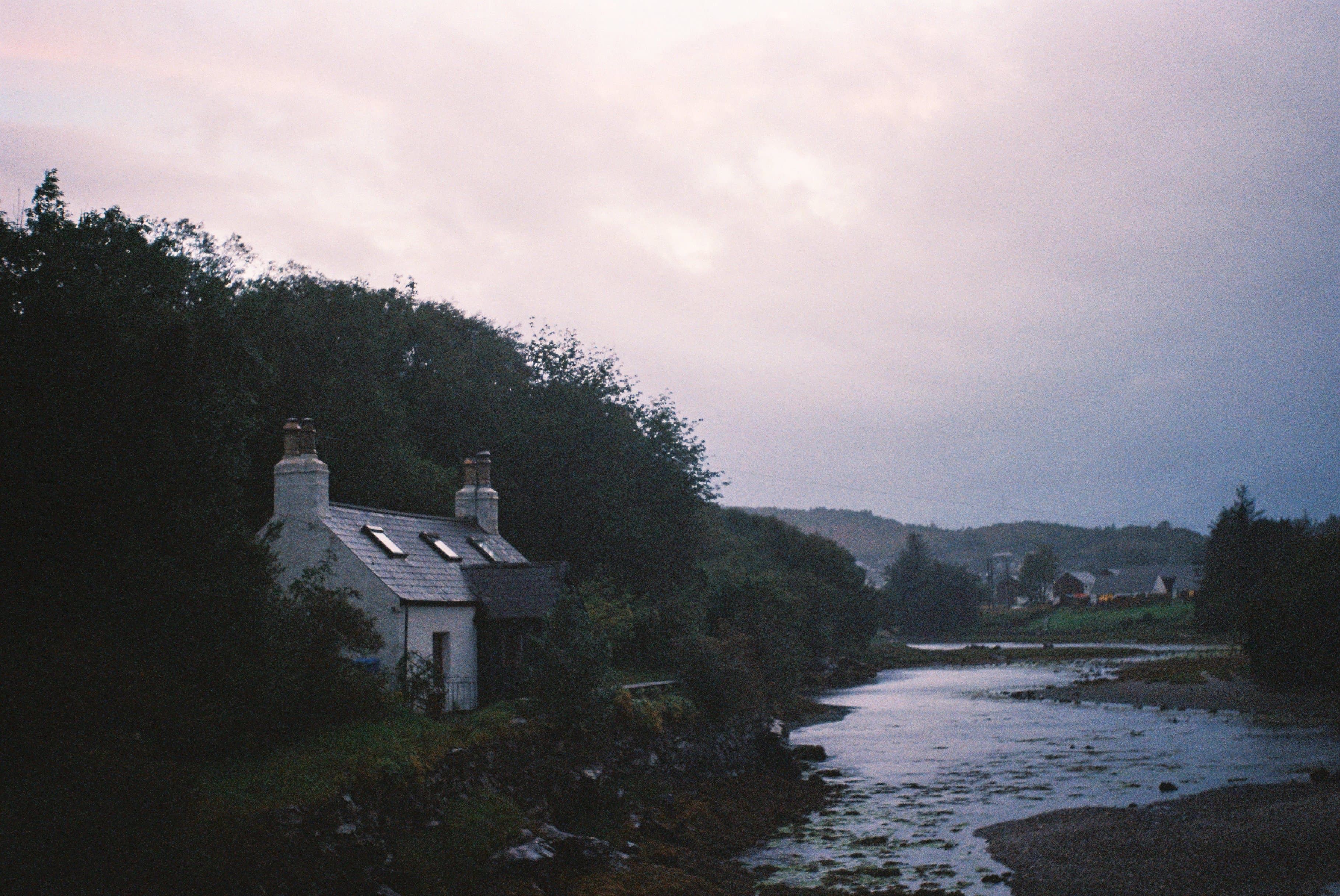 A small white cottage surrounded by trees sits on an elevated shorebank adjacent to a river.