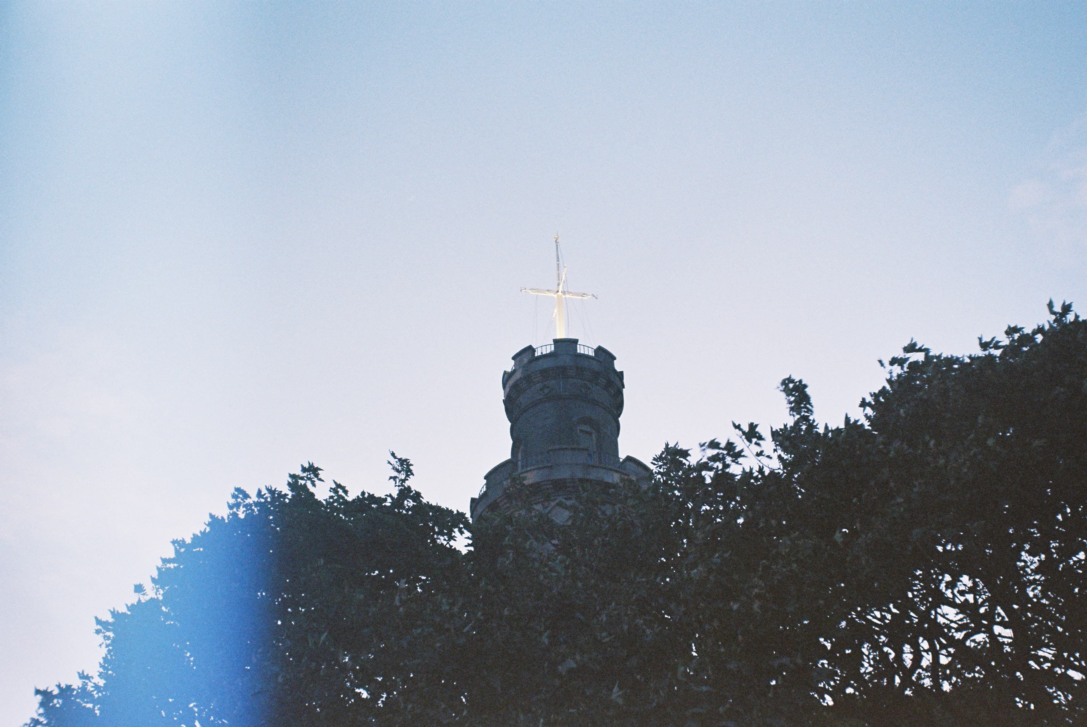 A tall, thin stone tower with a white cross above it. The base of the tower is covered by trees in the foreground. There is a film light leak visible on the left side of the image.