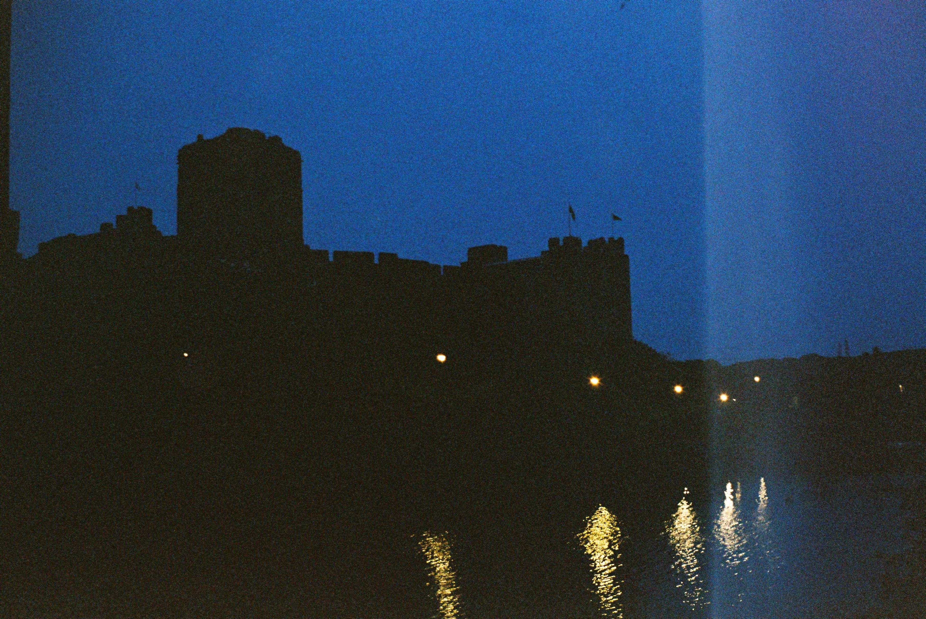 A silhouette of a castle against a deep blue sky. The light of five streetlamps are visible, with their light reflected in the water in the foreground. There is a film light leak on the right side of the image.